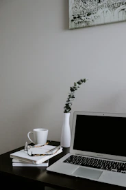 A minimalist workspace featuring a laptop, a notebook, and a cup of coffee beside an open book.