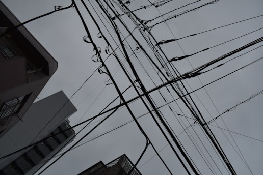 A complex network of black power lines crisscross overhead against a backdrop of a cloudy, grey sky. Buildings rise up from the bottom edges of the image, framing the scene and adding an urban feel.