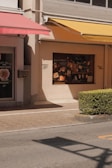 A street scene featuring the facade of a building with two colorful awnings, one red and one yellow, casting shadows on the wall. The display window showcases various posters and images related to food and drinks. A neatly trimmed bush lines the edge of a pavement, with the street in the foreground.