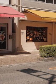A street scene featuring the facade of a building with two colorful awnings, one red and one yellow, casting shadows on the wall. The display window showcases various posters and images related to food and drinks. A neatly trimmed bush lines the edge of a pavement, with the street in the foreground.