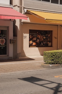 A street scene featuring the facade of a building with two colorful awnings, one red and one yellow, casting shadows on the wall. The display window showcases various posters and images related to food and drinks. A neatly trimmed bush lines the edge of a pavement, with the street in the foreground.