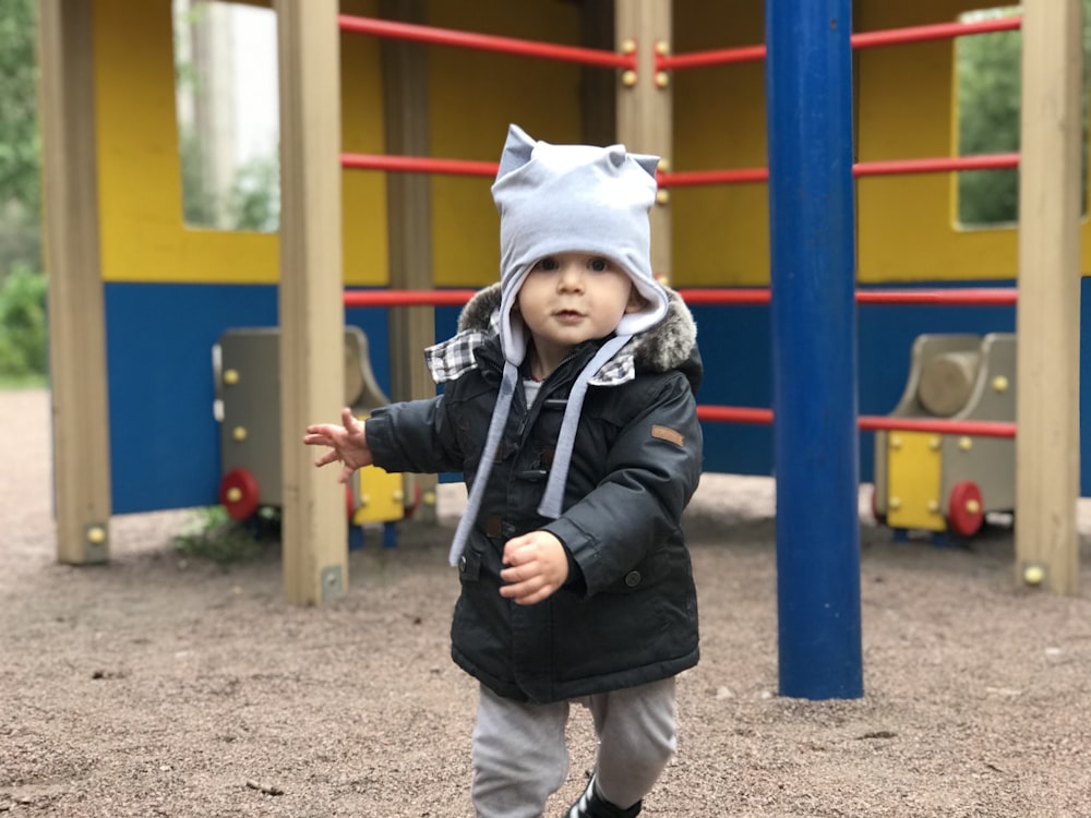 niño con chaqueta negra y gorra blanca de pie cerca del patio de recreo amarillo, azul y rojo durante el día