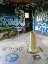 A small room covered in colorful graffiti on the walls, ceiling, and floor. There are various spray-painted designs and words, with a yellow post in the middle of the floor. A concrete bench along the curved wall is also marked with graffiti. Light comes in through an open doorway leading to another room.