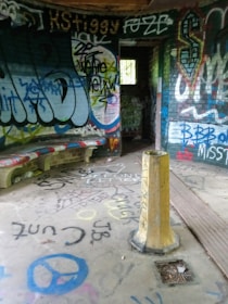 A small room covered in colorful graffiti on the walls, ceiling, and floor. There are various spray-painted designs and words, with a yellow post in the middle of the floor. A concrete bench along the curved wall is also marked with graffiti. Light comes in through an open doorway leading to another room.
