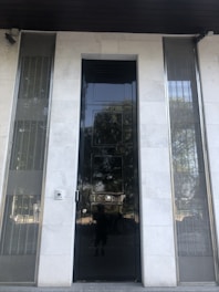 A tall, glossy black door is centered on a facade made of light-colored stone. The door has a reflective surface, with visible distortions and reflections. On either side of the door, there are vertical metal grilles set within the stone facade. A small security camera is mounted above the door, and a doorbell or intercom is situated to the left of the door.