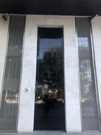 A tall, glossy black door is centered on a facade made of light-colored stone. The door has a reflective surface, with visible distortions and reflections. On either side of the door, there are vertical metal grilles set within the stone facade. A small security camera is mounted above the door, and a doorbell or intercom is situated to the left of the door.