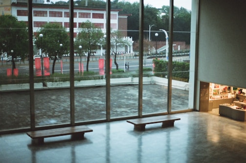 A spacious, modern interior with large floor-to-ceiling windows overlooking a street and red banners outside. Two wooden benches are placed facing the windows, and a small shop area is visible on the right, displaying various items. Green trees and a building can be seen through the windows.