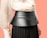 A close-up view of a person wearing a fashionable outfit, featuring a structured black belt or waistband made of leather with zipper details. The top is a light cream blouse with sheer, flowing sleeves, and the skirt is dark pleated fabric. The background is a solid peach color.