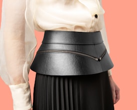 A close-up view of a person wearing a fashionable outfit, featuring a structured black belt or waistband made of leather with zipper details. The top is a light cream blouse with sheer, flowing sleeves, and the skirt is dark pleated fabric. The background is a solid peach color.
