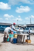 Two people are gathering water or some liquid into buckets using a makeshift setup that includes a corrugated metal sheet and various colorful plastic containers. They are in an outdoor setting with cars parked in the background, and a somewhat rural environment is suggested by the surrounding structures.