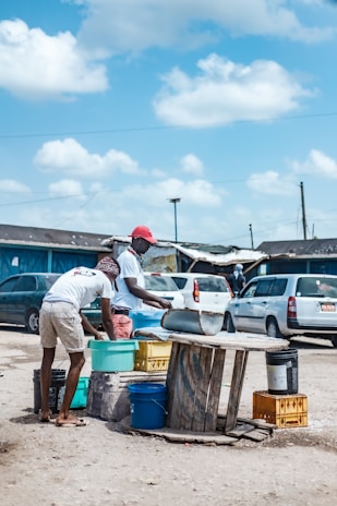 Two people are gathering water or some liquid into buckets using a makeshift setup that includes a corrugated metal sheet and various colorful plastic containers. They are in an outdoor setting with cars parked in the background, and a somewhat rural environment is suggested by the surrounding structures.