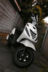 A white scooter is parked next to a wooden fence on a paved surface. The scooter appears clean and well-maintained, with a focus on its front tire and handlebars. Surrounding the area are wooden panels and leafy vegetation, suggesting an outdoor setting.