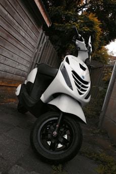 A white scooter is parked next to a wooden fence on a paved surface. The scooter appears clean and well-maintained, with a focus on its front tire and handlebars. Surrounding the area are wooden panels and leafy vegetation, suggesting an outdoor setting.