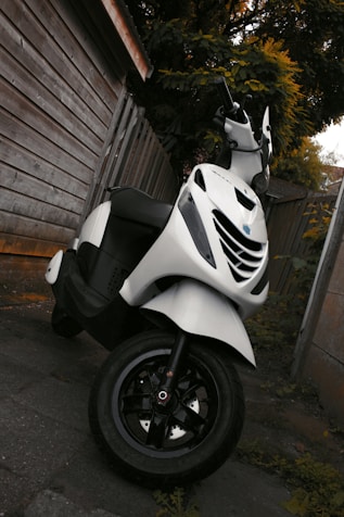 A white scooter is parked next to a wooden fence on a paved surface. The scooter appears clean and well-maintained, with a focus on its front tire and handlebars. Surrounding the area are wooden panels and leafy vegetation, suggesting an outdoor setting.
