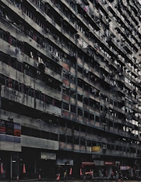 A large, aged, and densely populated apartment building with multiple floors, characterized by small balconies and air conditioning units attached to windows. The lower part of the building includes various shops and businesses, with signs in another language. Traffic cones and red flags are visible at the sidewalk. The building has a gritty, worn-out appearance.