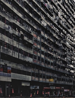 A large, aged, and densely populated apartment building with multiple floors, characterized by small balconies and air conditioning units attached to windows. The lower part of the building includes various shops and businesses, with signs in another language. Traffic cones and red flags are visible at the sidewalk. The building has a gritty, worn-out appearance.