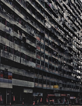 A large, aged, and densely populated apartment building with multiple floors, characterized by small balconies and air conditioning units attached to windows. The lower part of the building includes various shops and businesses, with signs in another language. Traffic cones and red flags are visible at the sidewalk. The building has a gritty, worn-out appearance.