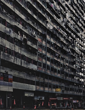 A large, aged, and densely populated apartment building with multiple floors, characterized by small balconies and air conditioning units attached to windows. The lower part of the building includes various shops and businesses, with signs in another language. Traffic cones and red flags are visible at the sidewalk. The building has a gritty, worn-out appearance.