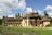 A charming, rustic house with thatched roofs and beige stone walls, surrounded by lush greenery and set beside a small pond. Several people are walking around the building, and the sky is a vivid blue with scattered clouds.
