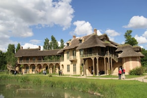 A charming, rustic house with thatched roofs and beige stone walls, surrounded by lush greenery and set beside a small pond. Several people are walking around the building, and the sky is a vivid blue with scattered clouds.