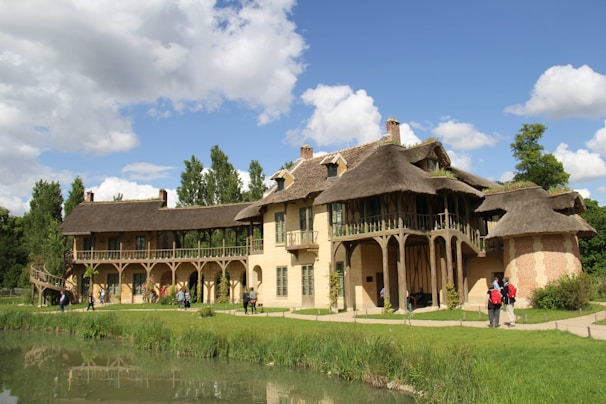 A charming, rustic house with thatched roofs and beige stone walls, surrounded by lush greenery and set beside a small pond. Several people are walking around the building, and the sky is a vivid blue with scattered clouds.