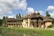 A charming, rustic house with thatched roofs and beige stone walls, surrounded by lush greenery and set beside a small pond. Several people are walking around the building, and the sky is a vivid blue with scattered clouds.