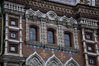 An ornate architectural facade featuring intricate details, with rows of arched windows framed by elaborate decorative elements. The facade is adorned with colored tiles and textures, depicting a mix of geometric and floral patterns. The building showcases richly detailed masonry with a combination of white stone and reddish-brown brick, creating a visually striking contrast.