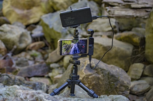 A smartphone mounted on a small tripod records a video of a waterfall in a rocky outdoor setting. The phone is connected to an external microphone through a cable. The background consists of large, uneven stones and natural elements, indicating a nature setting.