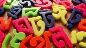 A collection of colorful, plastic Bengali alphabet letters scattered randomly across a brown surface. The letters feature vibrant shades of pink, green, blue, yellow, and orange, creating a lively and playful arrangement.