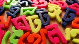 A collection of colorful, plastic Bengali alphabet letters scattered randomly across a brown surface. The letters feature vibrant shades of pink, green, blue, yellow, and orange, creating a lively and playful arrangement.