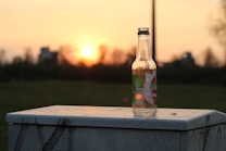 A glass bottle with a colorful label stands on a white, slightly weathered, outdoor surface. The background features a soft-focus sunset with a warm orange glow, casting a reflective light through the bottle, while dark silhouettes of trees and distant buildings are visible beneath the colorful sky.