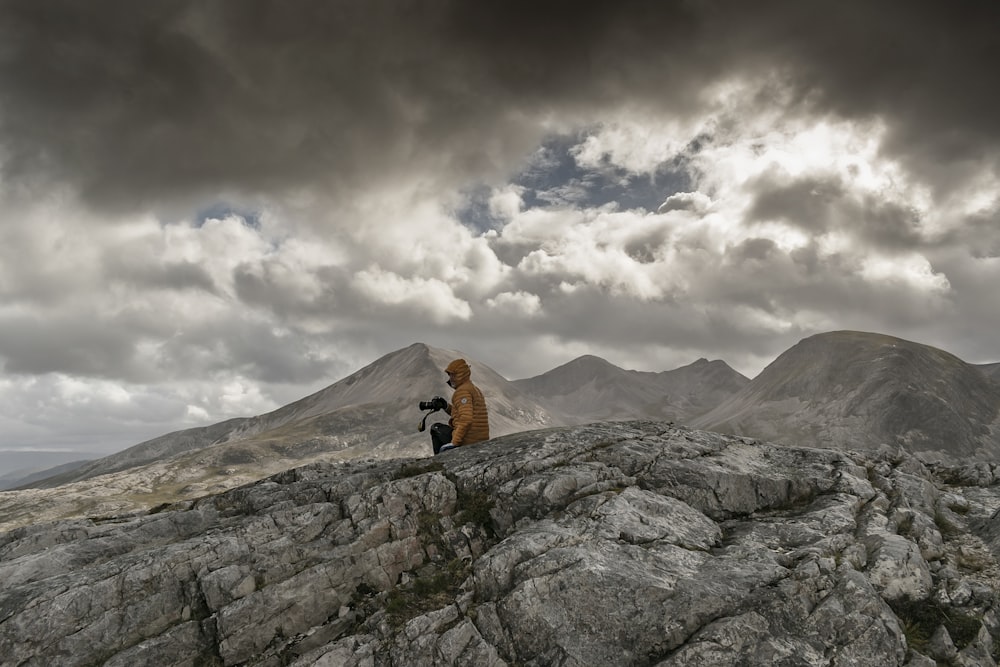 person in orange jacket sitting on rock mountain under white clouds during daytime