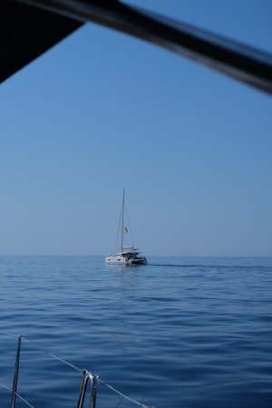 A sailboat glides across a calm, expansive body of water under a clear blue sky. The image captures the serene atmosphere of open waters on a sunny day, framed by parts of another boat in the foreground.