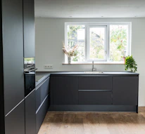 A modern kitchen with sleek, dark cabinetry and a minimalistic design. The kitchen features a large window allowing natural light to illuminate the space, with potted plants on the windowsill. The floor is wooden and contrasts with the dark cabinets, while the countertop has a built-in sink and faucet.