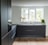 A modern kitchen with sleek, dark cabinetry and a minimalistic design. The kitchen features a large window allowing natural light to illuminate the space, with potted plants on the windowsill. The floor is wooden and contrasts with the dark cabinets, while the countertop has a built-in sink and faucet.