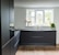 A modern kitchen with sleek, dark cabinetry and a minimalistic design. The kitchen features a large window allowing natural light to illuminate the space, with potted plants on the windowsill. The floor is wooden and contrasts with the dark cabinets, while the countertop has a built-in sink and faucet.