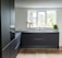 A modern kitchen with sleek, dark cabinetry and a minimalistic design. The kitchen features a large window allowing natural light to illuminate the space, with potted plants on the windowsill. The floor is wooden and contrasts with the dark cabinets, while the countertop has a built-in sink and faucet.