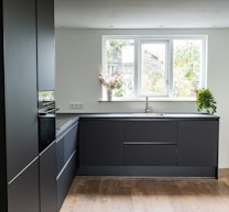A modern kitchen with sleek, dark cabinetry and a minimalistic design. The kitchen features a large window allowing natural light to illuminate the space, with potted plants on the windowsill. The floor is wooden and contrasts with the dark cabinets, while the countertop has a built-in sink and faucet.