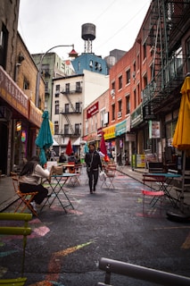 An urban street scene with colorful outdoor seating outside various shops and restaurants. People walk along the street, which is adorned with vibrant patterns. The buildings showcase a mix of old brick facades and eclectic signage, creating a lively urban atmosphere.