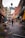 An urban street scene with colorful outdoor seating outside various shops and restaurants. People walk along the street, which is adorned with vibrant patterns. The buildings showcase a mix of old brick facades and eclectic signage, creating a lively urban atmosphere.