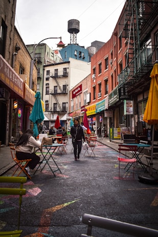An urban street scene with colorful outdoor seating outside various shops and restaurants. People walk along the street, which is adorned with vibrant patterns. The buildings showcase a mix of old brick facades and eclectic signage, creating a lively urban atmosphere.