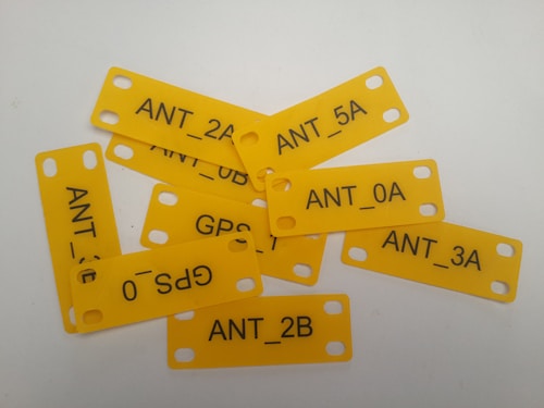 Several yellow rectangular tags with rounded corners are scattered on a flat surface. Each tag has two holes on either side and contains black text, showing combinations of letters and numbers such as GPS_0, ANT_5A, and ANT_0A.