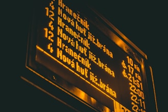 An illuminated electronic sign displaying bus or train schedules in yellow text against a dark background. The sign lists various destinations and times.