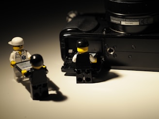 Three LEGO figures are interacting with a digital camera. One figure is wearing a white outfit and holding a piece of paper, while another figure is in black and appears to be assisting the third figure, who is accessing the open side of the camera.