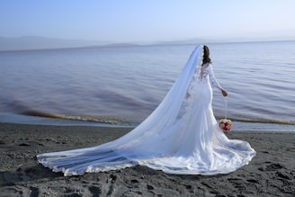 A bride in an elegant white wedding dress stands on a beach, facing the sea. She holds a colorful bouquet in one hand, and her long train flows behind her.