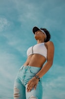 A confident person is looking up towards the sky, wearing a white sleeveless top and ripped blue jeans. The individual is sporting a visor cap and a watch on their wrist. The background is a clear blue sky.