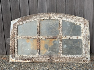 An antique, rectangular window frame with an arched top, featuring six glass panels. The frame is heavily rusted and worn, indicating significant age and exposure to the elements. The window is set against a wooden background, with the ground beneath being small stones.