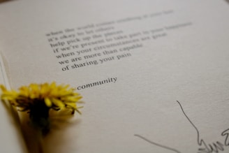 A close-up view of an open book with a yellow flower placed on the pages. The text on the page is partially visible, with the word 'community' clearly shown. The paper has a textured appearance, and there are minimal drawings near the edge.