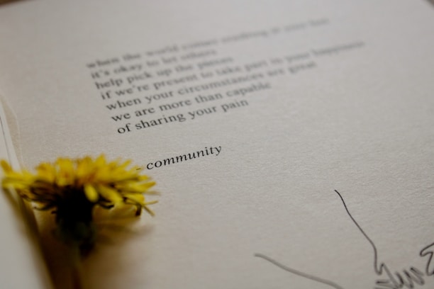 A close-up view of an open book with a yellow flower placed on the pages. The text on the page is partially visible, with the word 'community' clearly shown. The paper has a textured appearance, and there are minimal drawings near the edge.