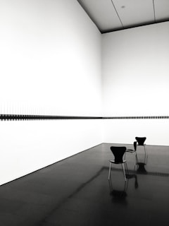 Two modern black chairs are placed in a white-walled room with a dark floor. The space features a minimalist art installation along the wall, composed of a series of aligned elements creating a monochromatic contrast.